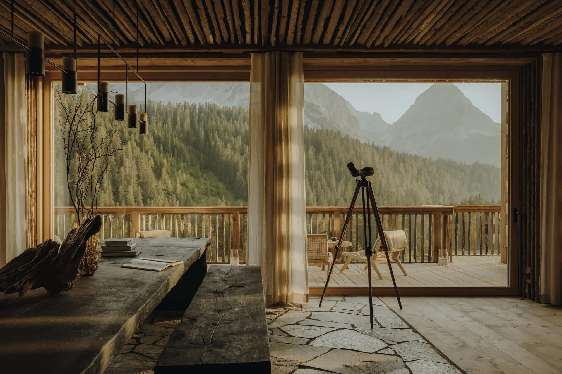A mountain view through the wide window of a timber chalet, a telescope standing on the stone floor.