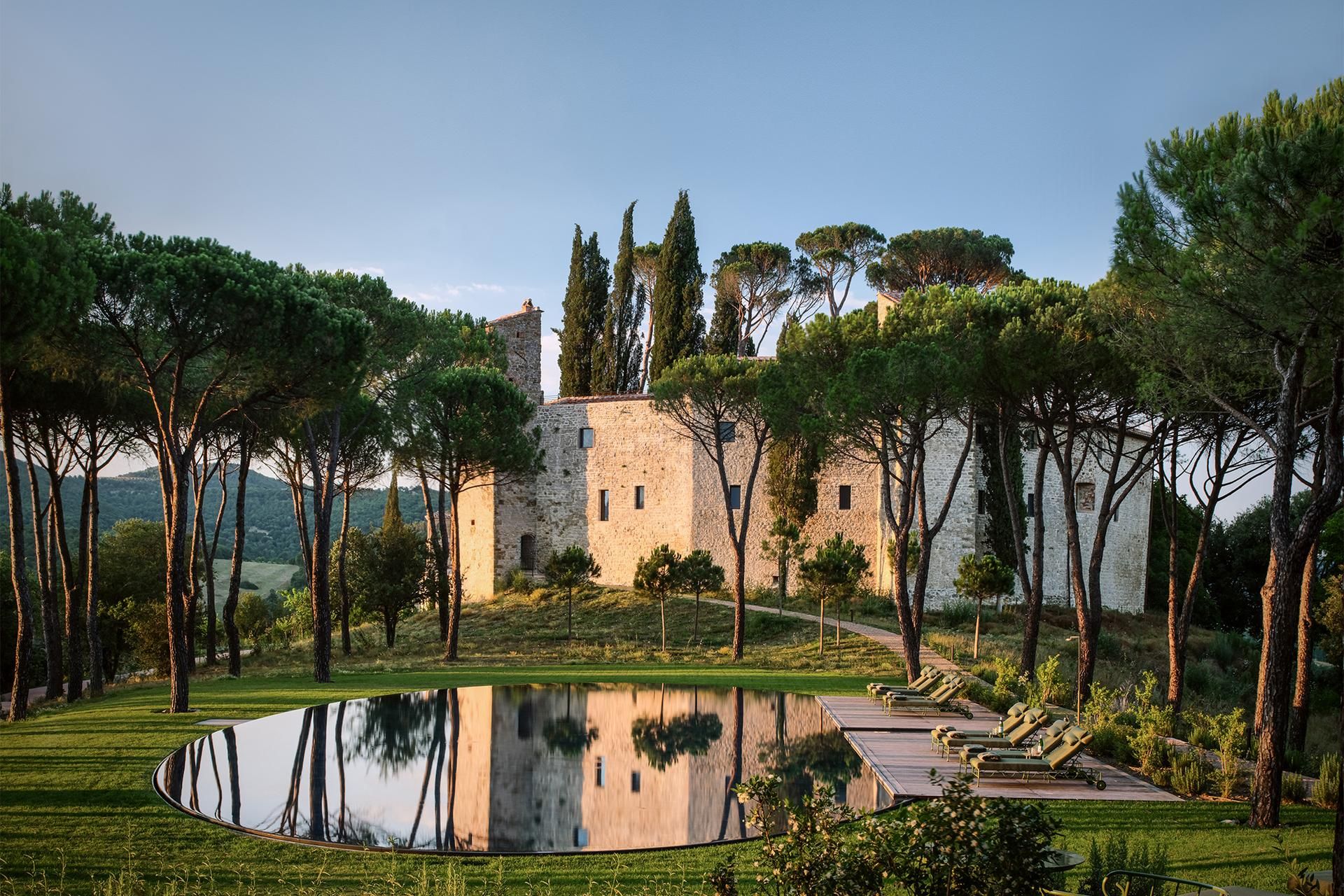 A stone castle among umbrella pines, reflected in a still circular pool in the evening light.
